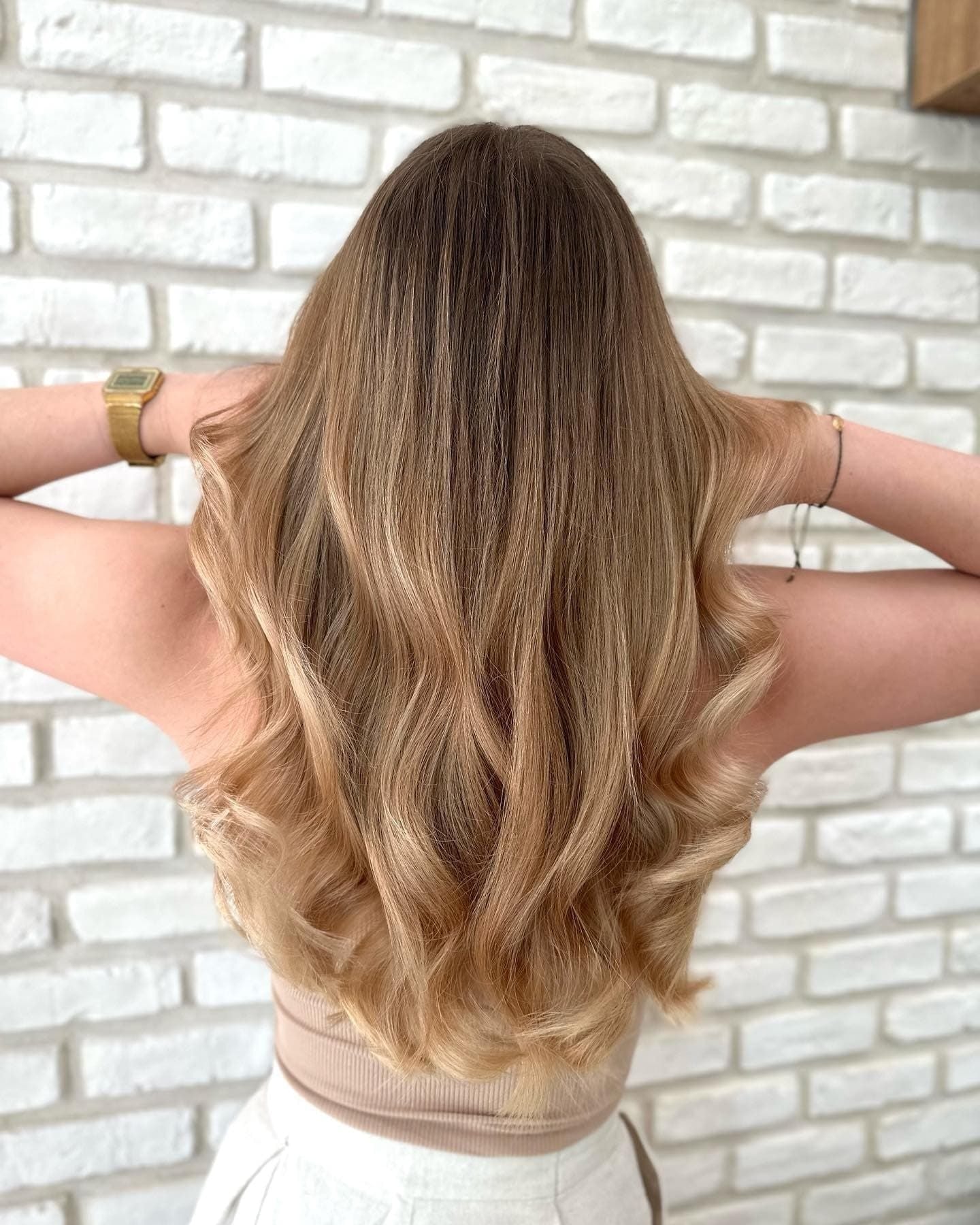 Balayage: dlouhé volné vlny v medové blond s tmavším kořínkem, ruce za hlavou