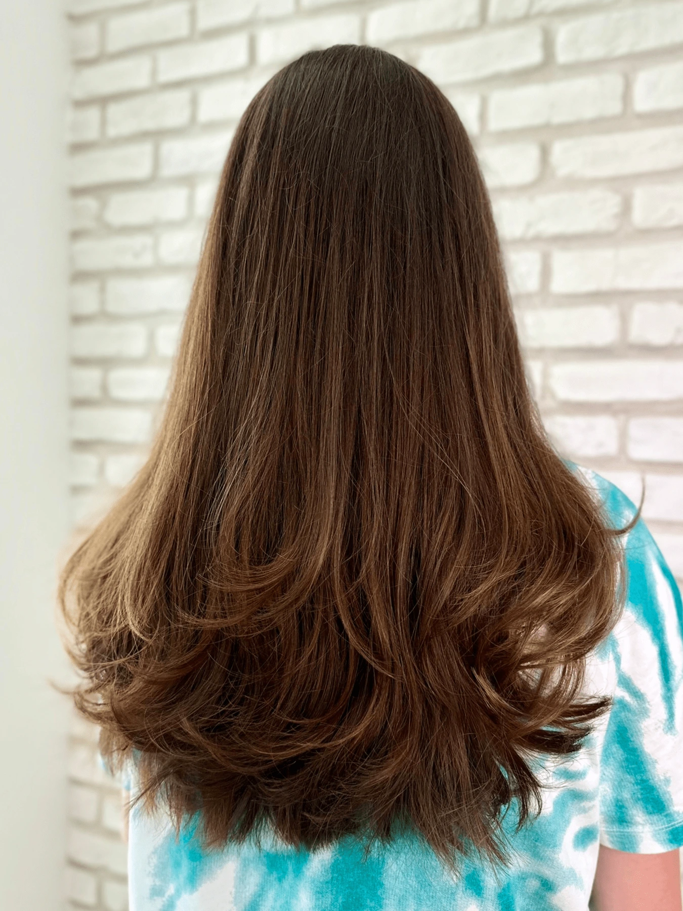 Butterfly cut: dlouhé hnědé vlasy s karamelovým nádechem, objemné vrstvy zvlněné na koncích, bílá cihlová stěna