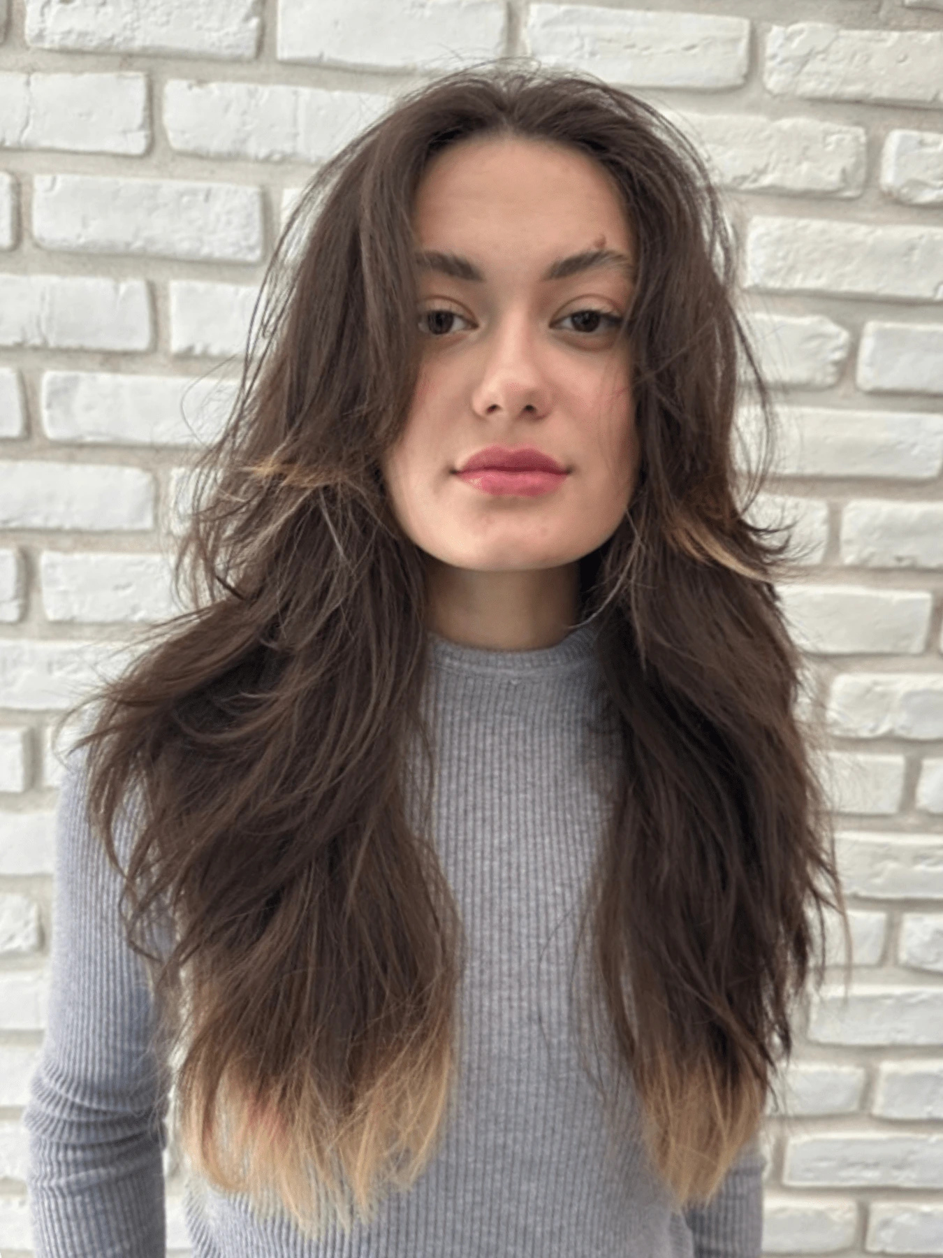 Butterfly cut: dlouhé tmavě hnědé vlasy s roztřepenými vrstvami, světlejší konečky, delší pramínky kolem obličeje