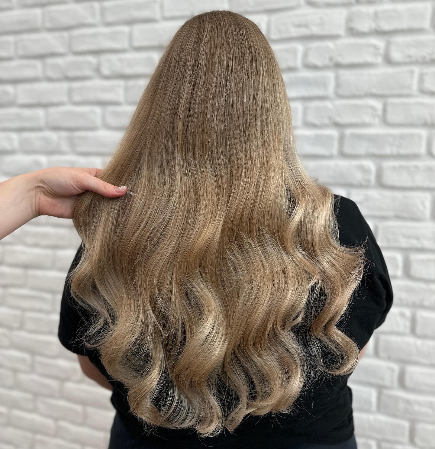 Kreativní barvení: dlouhé vlnité vlasy v medové blond s tmavším kořínkem, prameny drží ruka, bílá cihlová zeď (Sára Hedbávná)