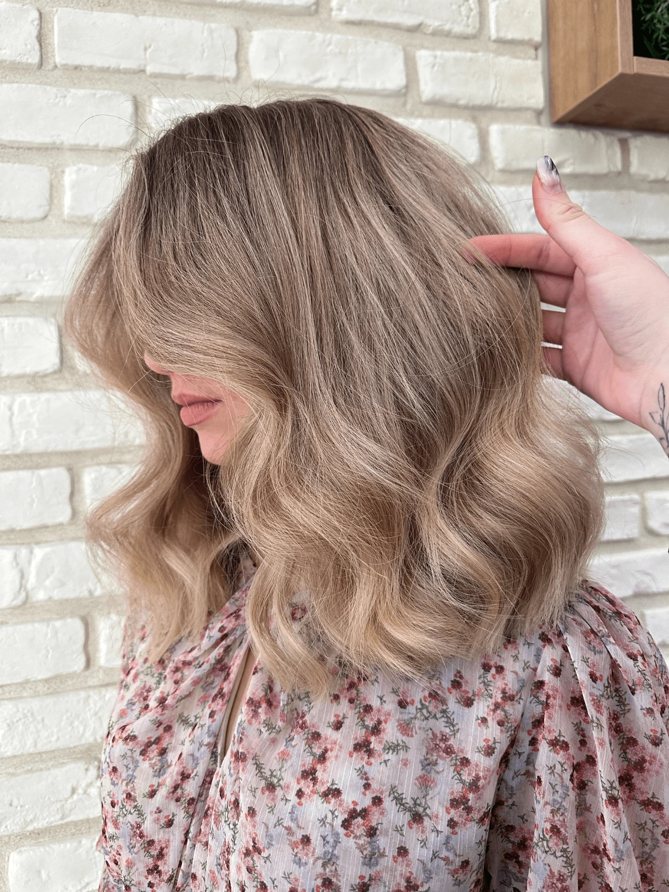 Foukaná: vlnité vlasy po ramena, písečná blond s tmavšími kořínky, objemný styling, květované oblečení