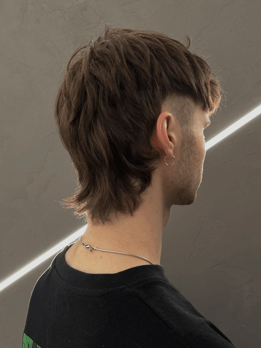 Mullet: hnědé stupňované vlasy delší vzadu, krátký zkosený bok, rozcuchaný styling, šedá zeď
