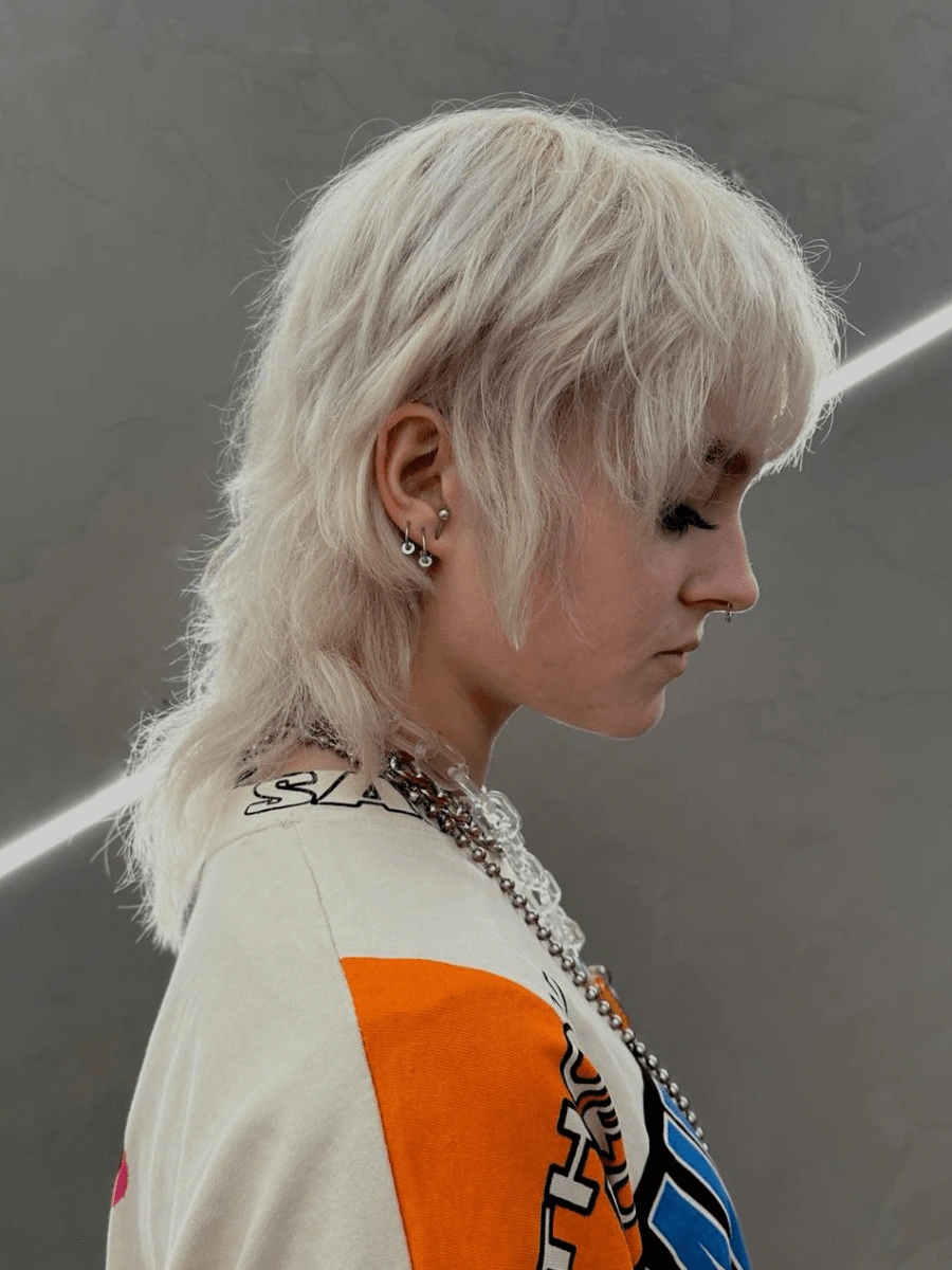 Střih vlasů: platinově blond rozcuchané vrstvené vlasy s ofinou, delší vzadu, šedé pozadí