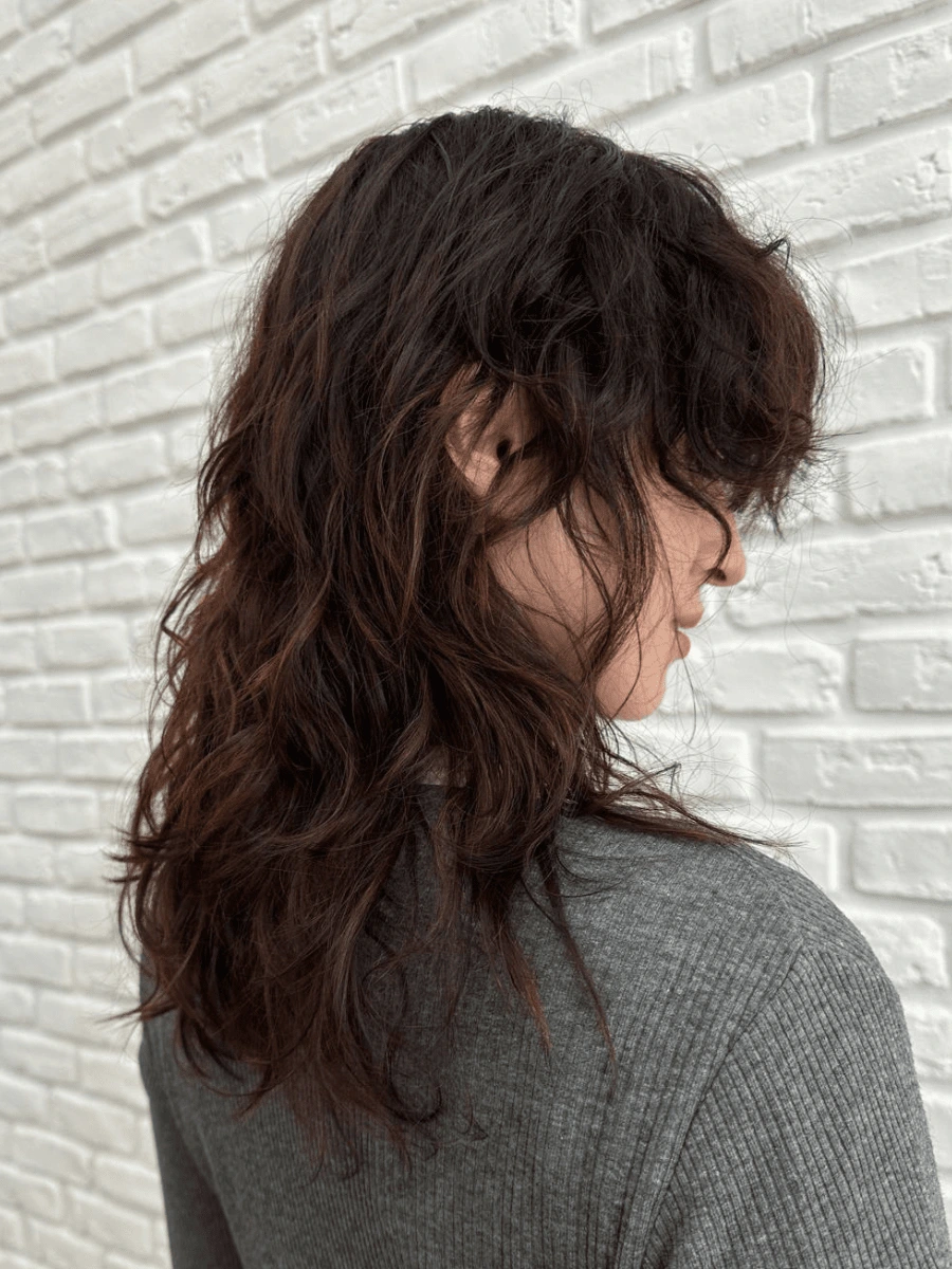Mullet: polodlouhé tmavě hnědé vlnité vlasy s ofinou do obličeje a stupňovanými délkami, bílá cihlová zeď
