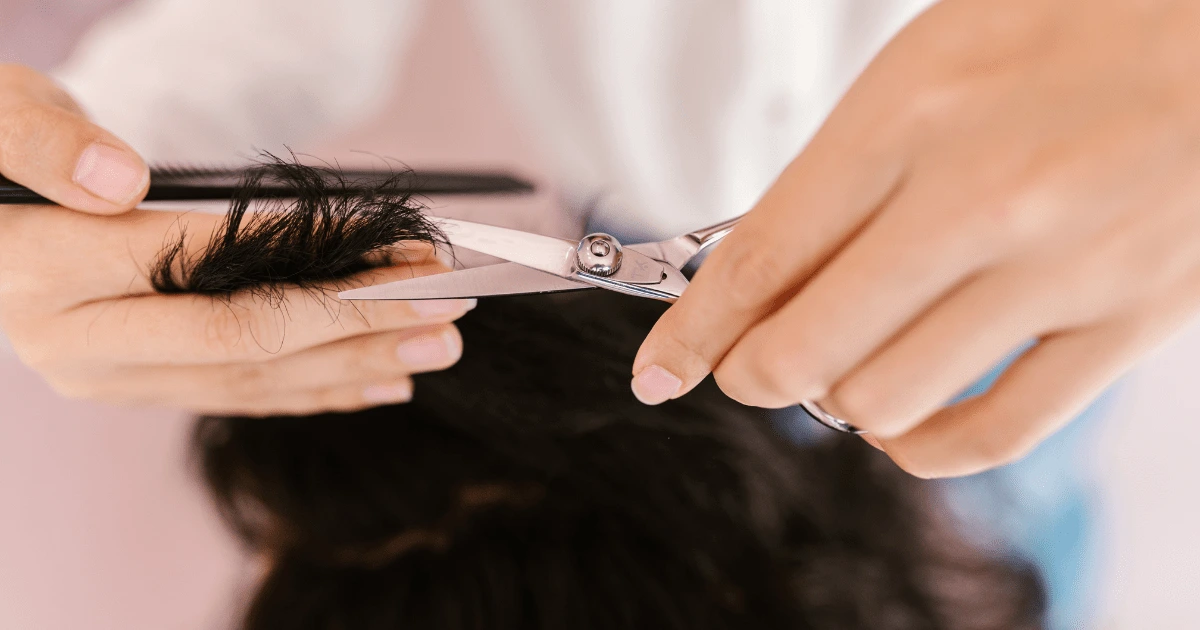 Mullet: ruce zastřihují nůžkami konečky tmavých vlnitých vlasů, druhá ruka drží pramen s hřebenem, růžové pozadí