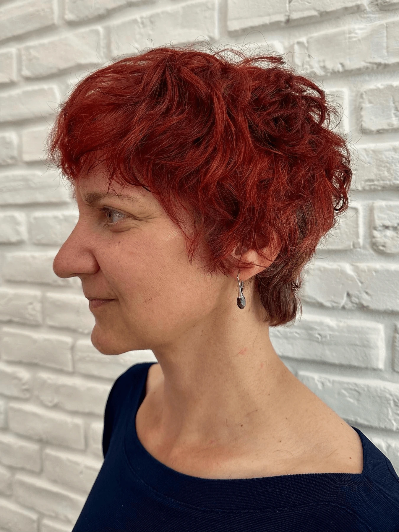 Pixie cut: krátké sytě červené vlnité vlasy s ofinou a kratším zátylkem, bílá cihlová zeď