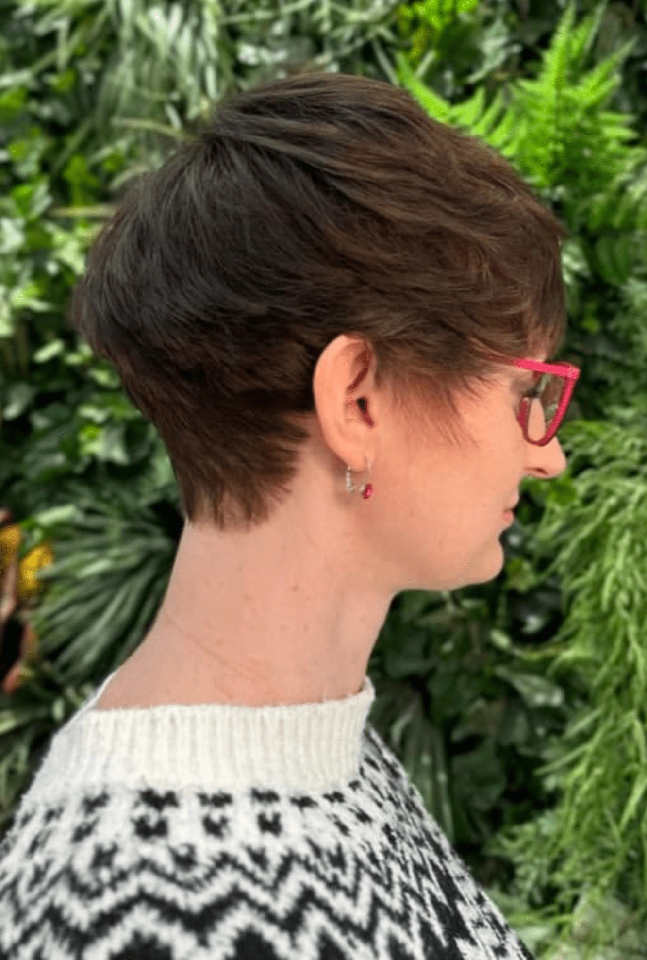 Pixie cut: krátké tmavě hnědé vlasy s jemnými světlejšími odlesky, delší nahoře, zelené pozadí