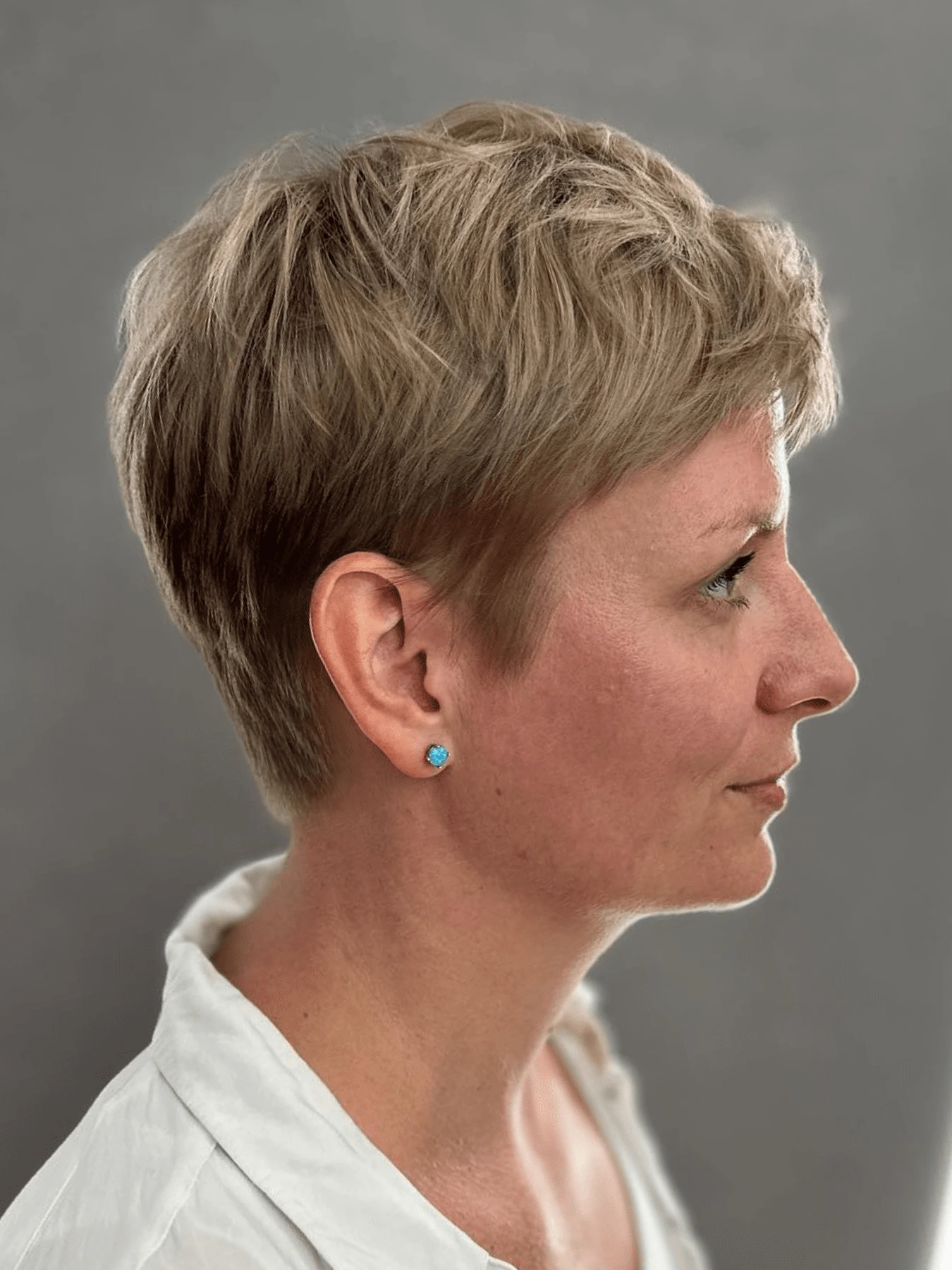 Pixie cut: velmi krátké vlasy v popelavém blond s texturovaným vrškem, krátký zátylek, šedé pozadí